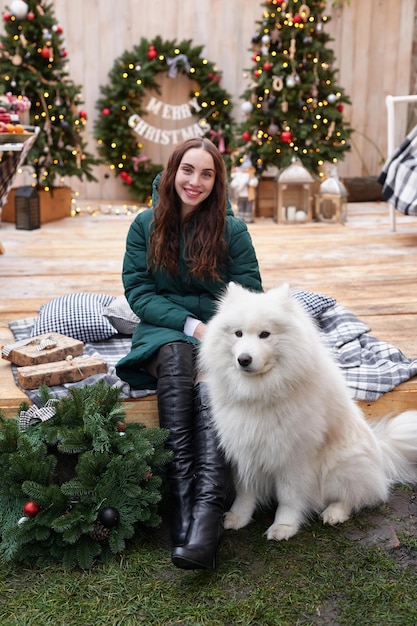 Giovane donna sullo sfondo dell'albero di Natale con il cane samoiedo bianco all'aperto Decorazione del cortile per il nuovo anno