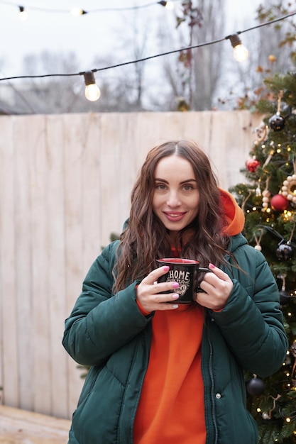 Giovane donna sullo sfondo dell'albero di Natale con una tazza di caffè all'aperto Decorazione del cortile capodanno