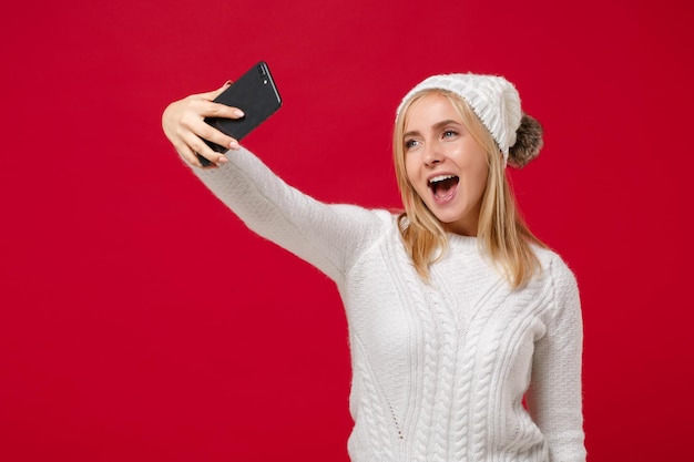 Giovane donna sveglia allegra in maglione bianco, cappello isolato sul ritratto dello studio del fondo della parete rossa. Stile di vita sano della moda, concetto di stagione fredda. Simula lo spazio della copia. Fare selfie girato sul cellulare.