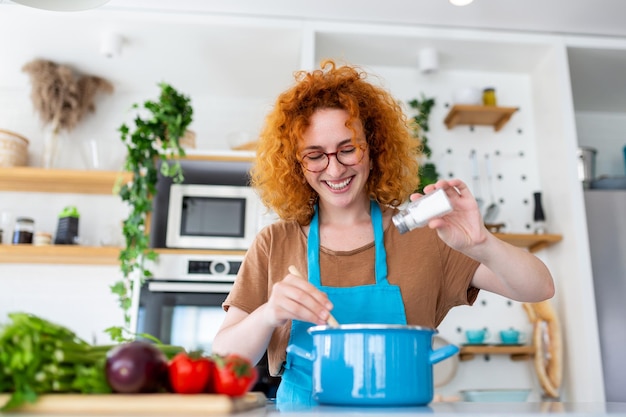 Giovane donna sveglia che cucina e aggiunge spezie al pasto ridendo e trascorrendo del tempo in cucina