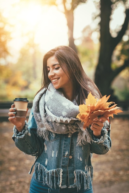 Giovane donna sveglia che gode nella foresta soleggiata nei colori di autunno. Ha in mano foglie gialle dorate e una tazza di caffè.