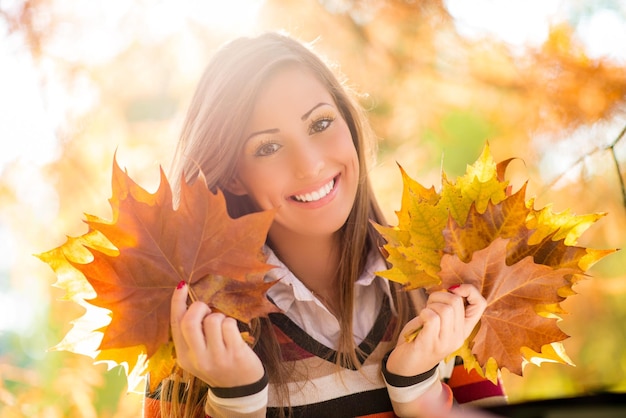 Giovane donna sveglia che gode nella foresta soleggiata nei colori di autunno. Ha in mano foglie giallo oro. Guardando la fotocamera.