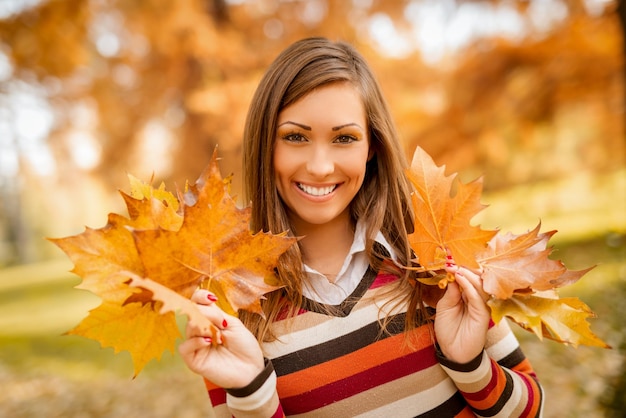 Giovane donna sveglia che gode nella foresta soleggiata nei colori di autunno. Sta tenendo foglie gialle dorate e sta guardando la fotocamera.