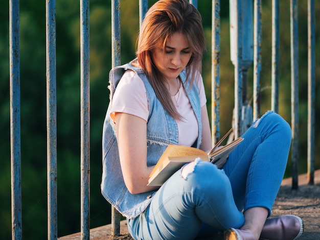 Giovane donna sveglia che legge un libro che si siede su un ponte