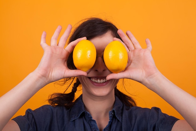 Giovane donna sveglia che sorride alla macchina fotografica con le arance come occhi sopra fondo giallo in studio. Arance succose. Arance gustose. Signora con gustose arance.