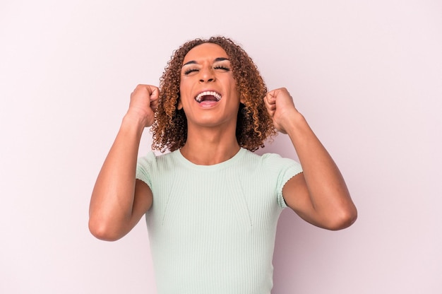 Giovane donna transessuale latina isolata su sfondo rosa che celebra una vittoria, passione ed entusiasmo, espressione felice.