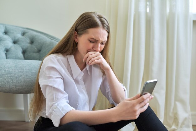 Giovane donna triste che piange con lo smartphone seduto sul pavimento a casa