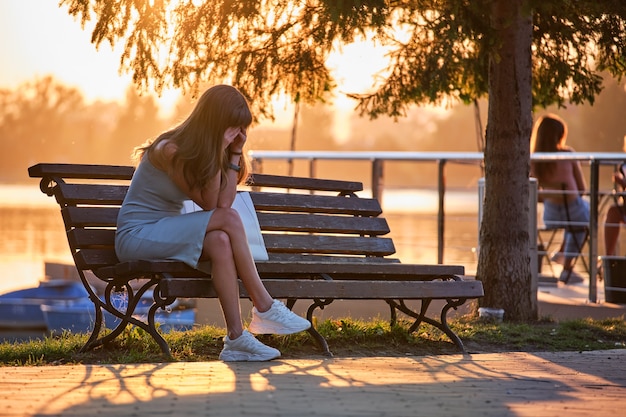 Giovane donna triste in abito estivo seduto da solo su una panchina sul lato del lago in una calda serata. Solitudine e riposo dal concetto di fretta quotidiana.