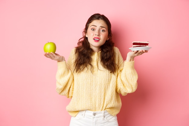 Giovane donna triste indecisa che scrolla le spalle che tiene mela sana verde e pezzo di torta di cibo spazzatura che sembra confuso e sconvolto in piedi contro il muro rosa