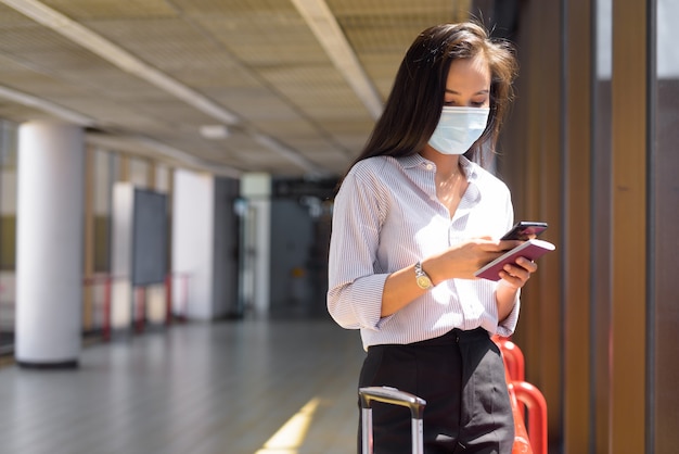 Giovane donna turistica asiatica con maschera utilizzando il telefono e tenendo il passaporto in aeroporto