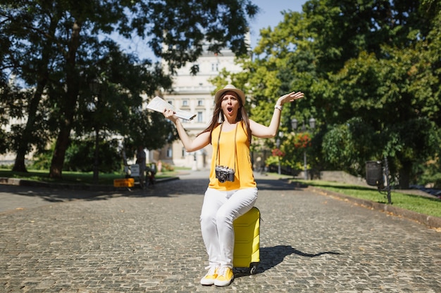 Giovane donna turistica irritata e arrabbiata del viaggiatore in vestiti gialli che si siede sulla mappa della città della tenuta della valigia che spande le mani all'aperto. Ragazza che viaggia all'estero per viaggiare nel fine settimana. Stile di vita del viaggio turistico.