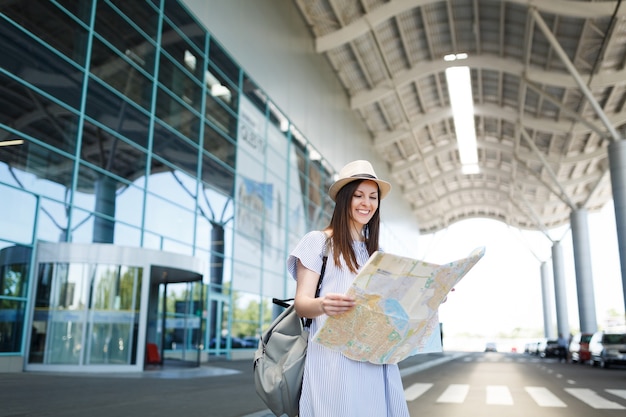 Giovane donna turistica sorridente del viaggiatore con lo zaino che tiene la mappa cartacea all'aeroporto internazionale