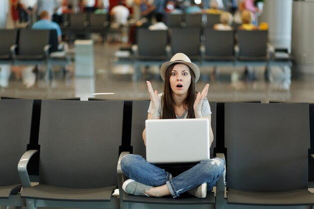 Giovane donna turistica viaggiatrice scioccata con laptop seduto con le gambe incrociate, allargando le mani mentre aspetta nella hall dell'aeroporto