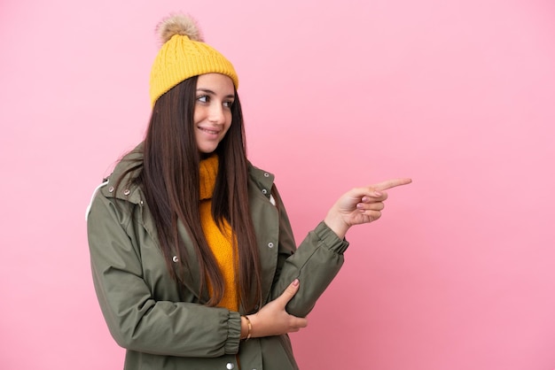 Giovane donna ucraina che indossa una giacca invernale isolata su sfondo rosa che punta il dito di lato e presenta un prodotto