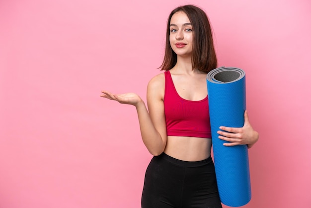 Giovane donna ucraina sportiva che va a lezioni di yoga mentre tiene un tappetino isolato su sfondo rosa che estende le mani di lato per invitare a venire