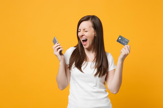 Giovane donna urlante pazza in abiti casual bianchi tenendo sì chiuso tenendo il telefono cellulare, carta di credito isolata su sfondo giallo arancione brillante. Concetto di stile di vita delle persone. Mock up spazio di copia.