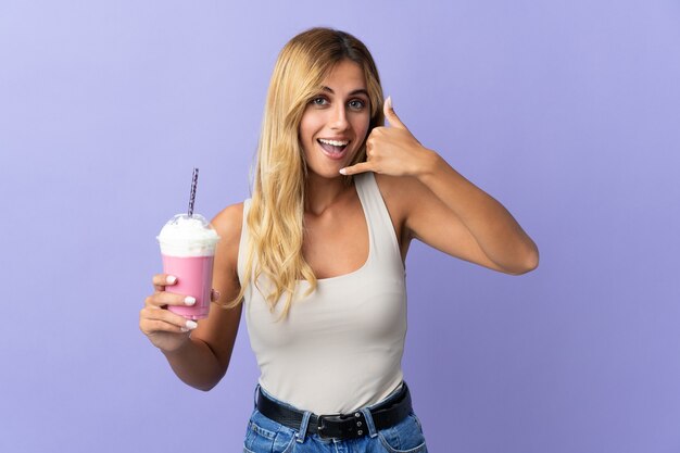 Giovane donna uruguaiana bionda con frappè alla fragola isolato sulla parete viola che fa gesto del telefono. Richiamami segno