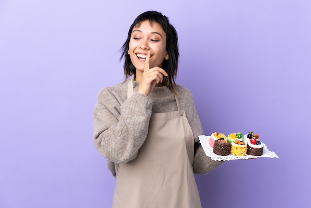 Giovane donna uruguaiana che tiene un sacco di mini torte diverse su viola isolato facendo il gesto del silenzio