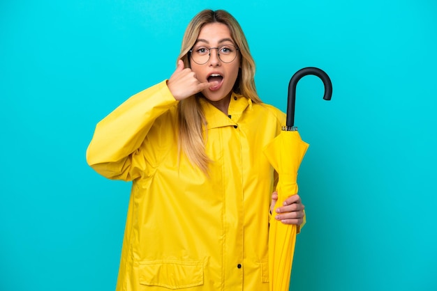 Giovane donna uruguaiana con cappotto antipioggia e ombrello isolato su sfondo blu che fa il gesto del telefono Call me back sign