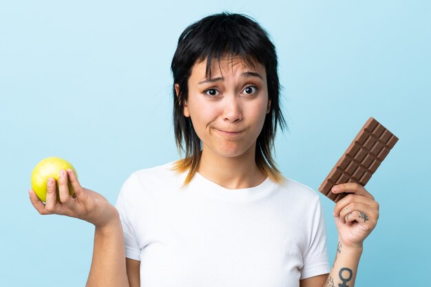 Giovane donna uruguaiana sopra la parete blu isolata che ha dubbi mentre prendendo una tavoletta di cioccolato in una mano e una mela nell'altra