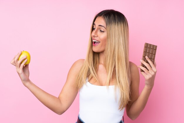 Giovane donna uruguaiana sul muro isolato rosa muro prendendo una tavoletta di cioccolato in una mano e una mela nell'altra