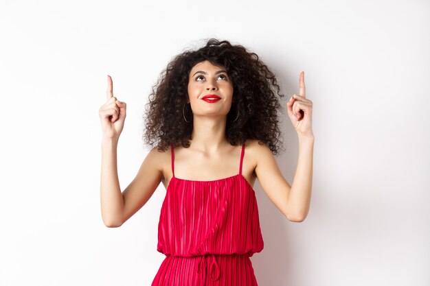Giovane donna vaga in vestito rosso alla moda, alzando lo sguardo e indicando il logo, sorridendo felice, in piedi su sfondo bianco. Copia spazio