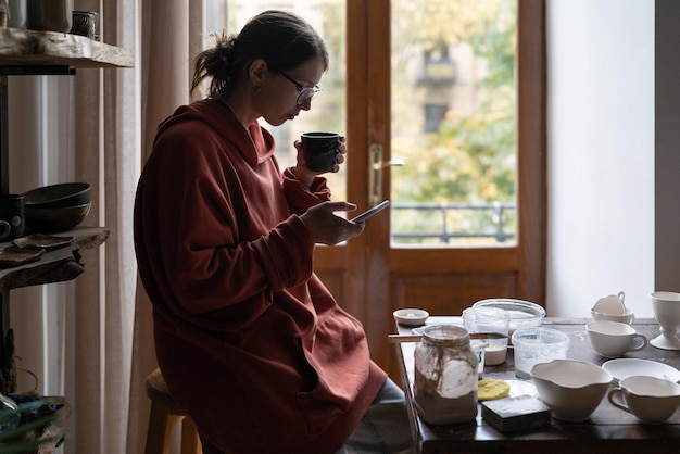 Giovane donna vasaio che riposa in un accogliente studio di ceramica domestica che beve tè e usa lo smartphone