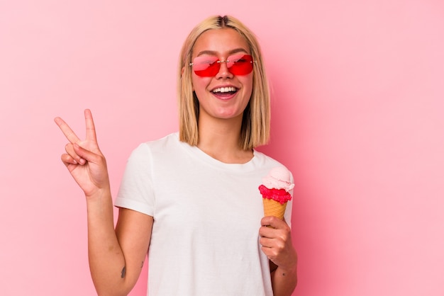 Giovane donna venezuelana che mangia un gelato isolato su sfondo rosa gioiosa e spensierata che mostra un simbolo di pace con le dita.