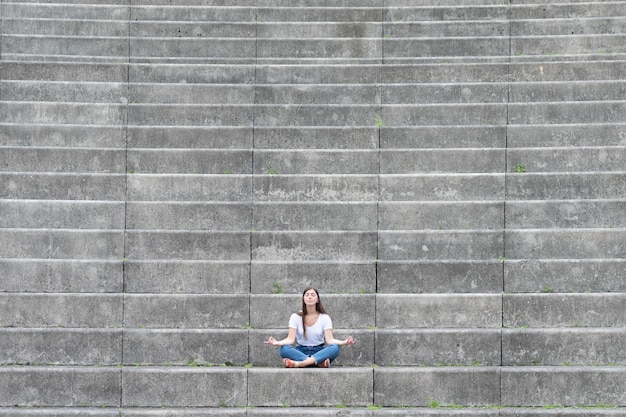 Giovane donna vestita casual seduta sulle scale di una tribuna e meditando. Salute mentale, consapevolezza e concetto di yoga con spazio di copia