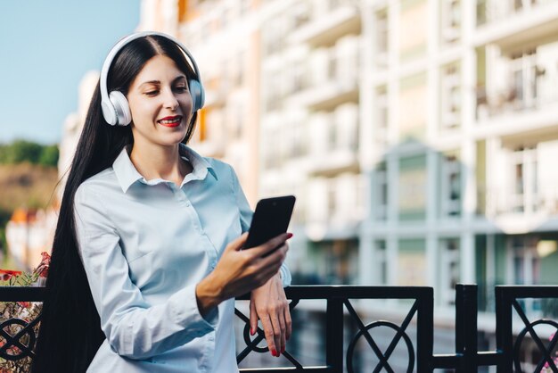 Giovane donna vestita casualmente sorridente graziosa in piedi sul balcone in via della città vecchia, godendo della vista e ascoltando musica sul suo telefono cellulare