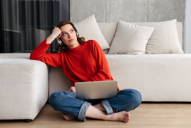 Giovane donna vestita casualmente stanca che si siede su un pavimento a casa, studiando con il computer portatile