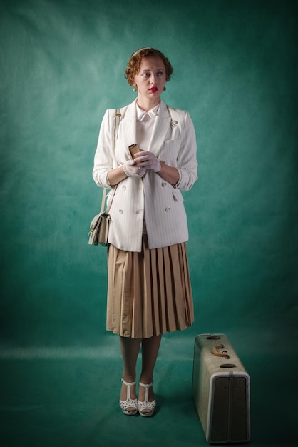 Giovane donna, vestita in stile retrò, con una valigia e un libro tra le mani. Ritratto in studio