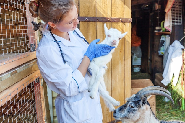 Giovane donna veterinaria con uno stetoscopio che tiene ed esamina un capretto sullo sfondo del ranch Giovane capretto nelle mani del veterinario per il controllo in una fattoria ecologica naturale Bestiame moderno allevamento ecologico