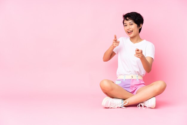Giovane donna vietnamita con i capelli corti che si siede sul pavimento sopra la parete rosa che indica e che sorride