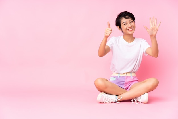 Giovane donna vietnamita con i capelli corti, seduta sul pavimento sul muro rosa contando sei con le dita