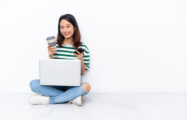 Giovane donna vietnamita con un computer portatile seduto sul pavimento isolato su un muro bianco che tiene il caffè da portare via e un cellulare