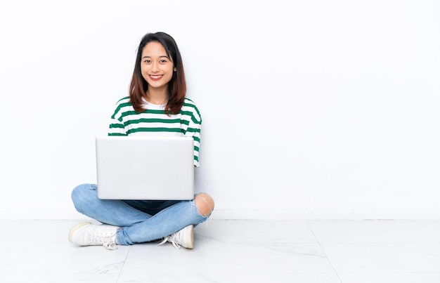 Giovane donna vietnamita con un computer portatile seduto sul pavimento isolato su un muro bianco tenendo le braccia incrociate in posizione frontale