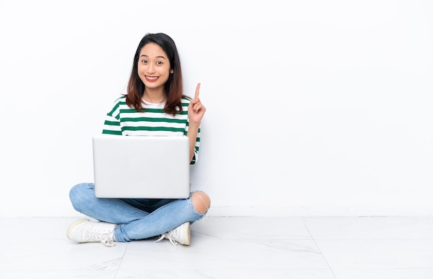 Giovane donna vietnamita con un computer portatile seduto sul pavimento isolato sul muro bianco che indica con il dito indice una grande idea