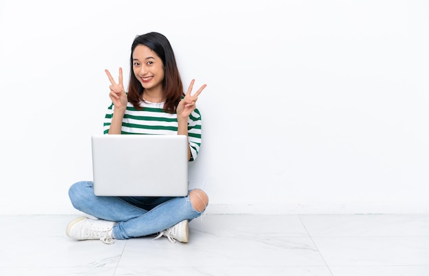 Giovane donna vietnamita con un computer portatile seduto sul pavimento isolato sul muro bianco che mostra il segno della vittoria con entrambe le mani