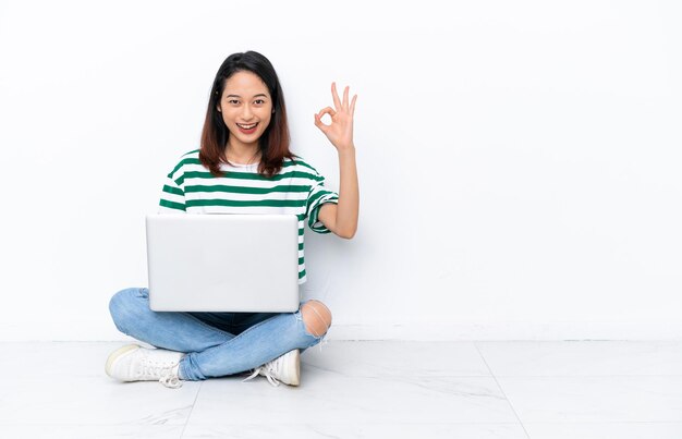 Giovane donna vietnamita con un computer portatile seduto sul pavimento isolato sul muro bianco che mostra segno ok con le dita