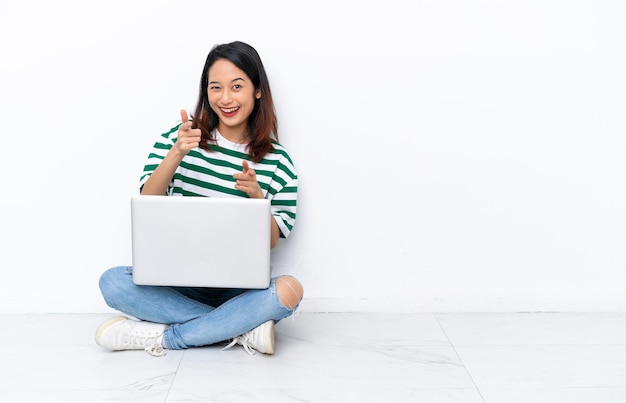 Giovane donna vietnamita con un computer portatile seduto sul pavimento isolato sul muro bianco che punta in avanti e sorridente