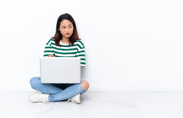 Giovane donna vietnamita con un laptop seduto sul pavimento isolato su un muro bianco che fa un gesto di dubbi sollevando le spalle