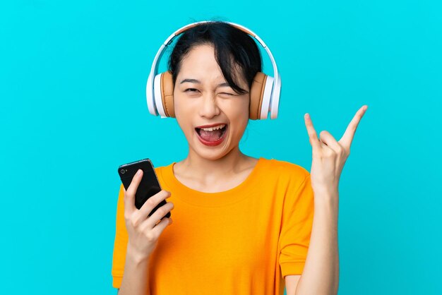Giovane donna vietnamita isolata su sfondo blu che ascolta musica con un cellulare che fa gesto rock
