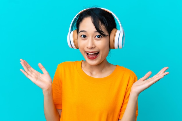 Giovane donna vietnamita isolata su sfondo blu sorpreso e ascoltando musica