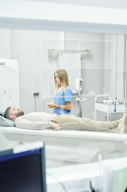 Giovane dottoressa bionda in uniforme medica in piedi da un paziente maschio barbuto sdraiato sulla cuccetta durante l'esame ecografico in cliniche