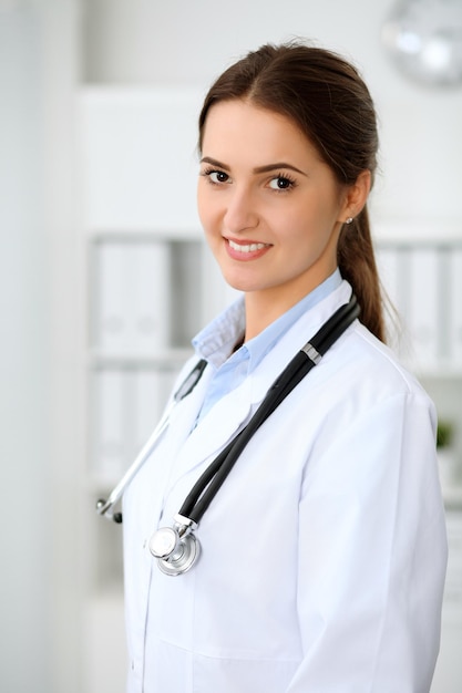 Giovane dottoressa bruna in piedi con appunti e sorridente in ospedale. Medico pronto ad esaminare il paziente
