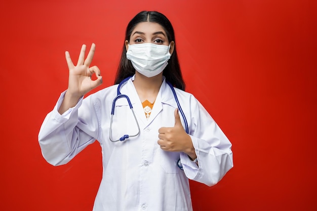 giovane dottoressa che indossa maschera, cappotto e stetoscopio su sfondo rosso modello pakistano indiano