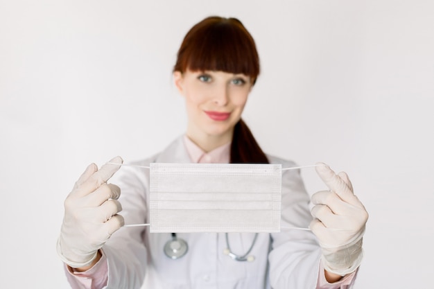 Giovane dottoressa con uno stetoscopio, in camice, sorridente e mostrando alla telecamera maschera protettiva contro le infezioni da virus, coved-19, in piedi su sfondo bianco. Focus sulla maschera nelle mani