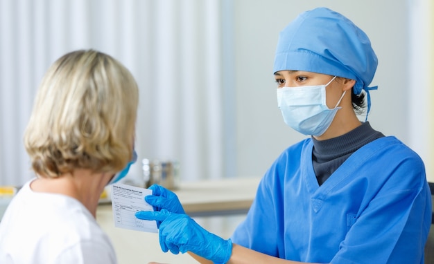 Giovane dottoressa in maschera facciale uniforme ospedaliera blu e guanti di gomma si siedono mostrando spiegazioni sulla carta di registrazione delle vaccinazioni Covid-19 certificano al paziente caucasico biondo.