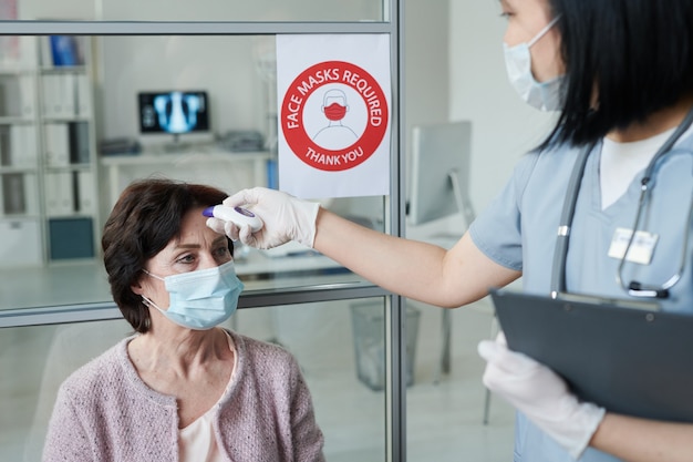 Giovane dottoressa o infermiera guantata in uniforme che misura la temperatura corporea del paziente maturo in maschera protettiva e abbigliamento casual